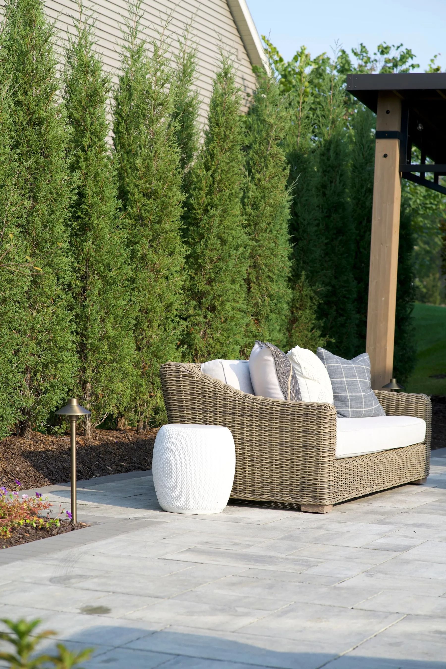Wicker sofa with cushions, white side table on a stone patio, with tall green trees in the background.