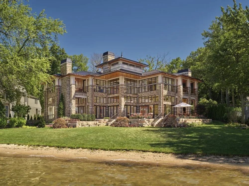 Multi-story stone house on a grassy bank near water, trees on either side, blue sky.