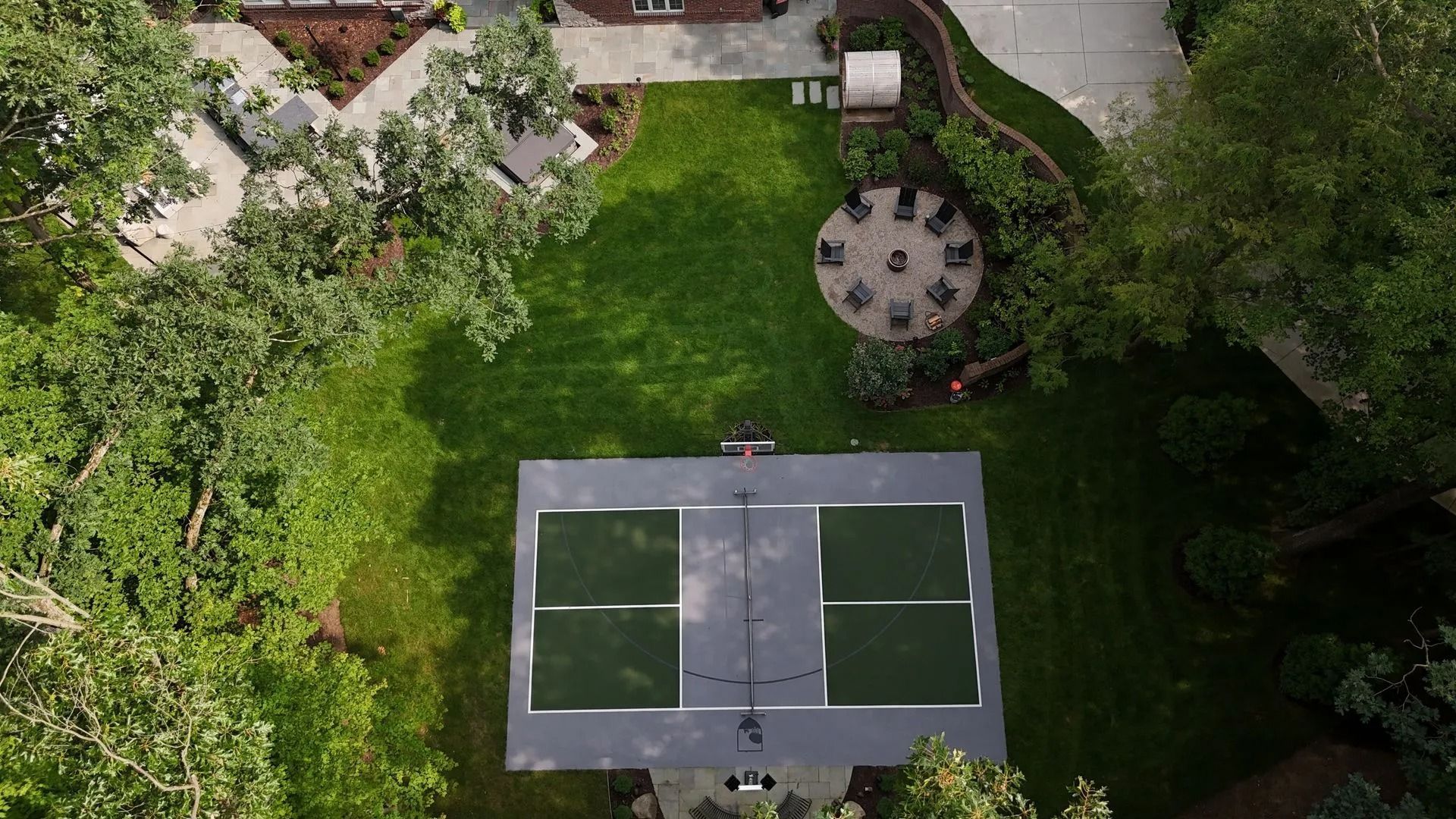 Aerial view: a backyard with a pickleball court, trees, and a patio with a round table and chairs.