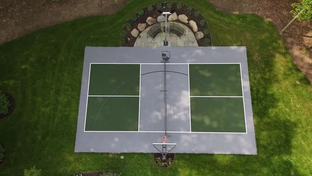 Overhead view of a dual-court sports area; grey concrete with green courts and basketball hoops, surrounded by grass and landscaping.