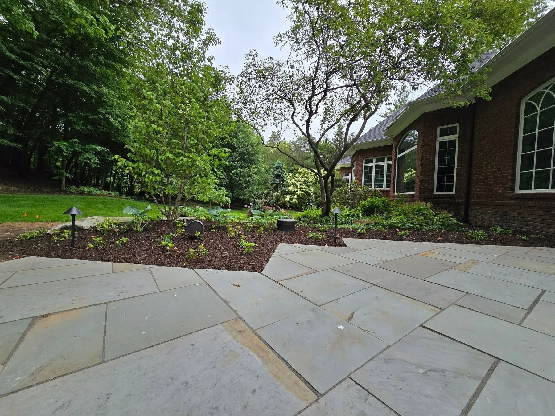 Stone patio with garden bed, trees, and a brick house. Overcast sky.