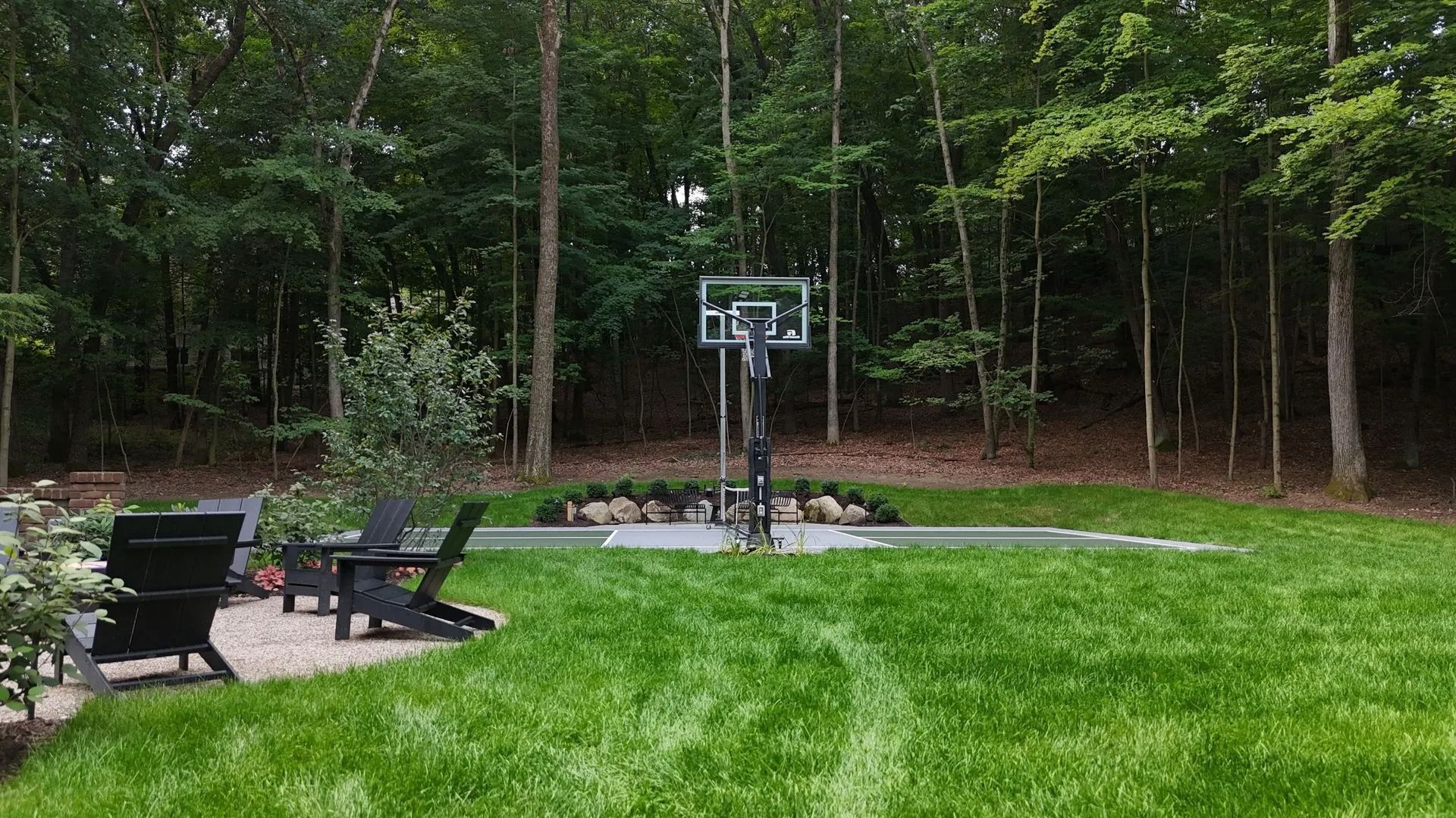 Backyard with basketball hoop, seating area, and lush green lawn surrounded by trees.