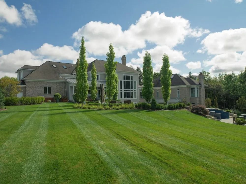 Large, brick house with a well-manicured lawn and tall trees under a partly cloudy sky.