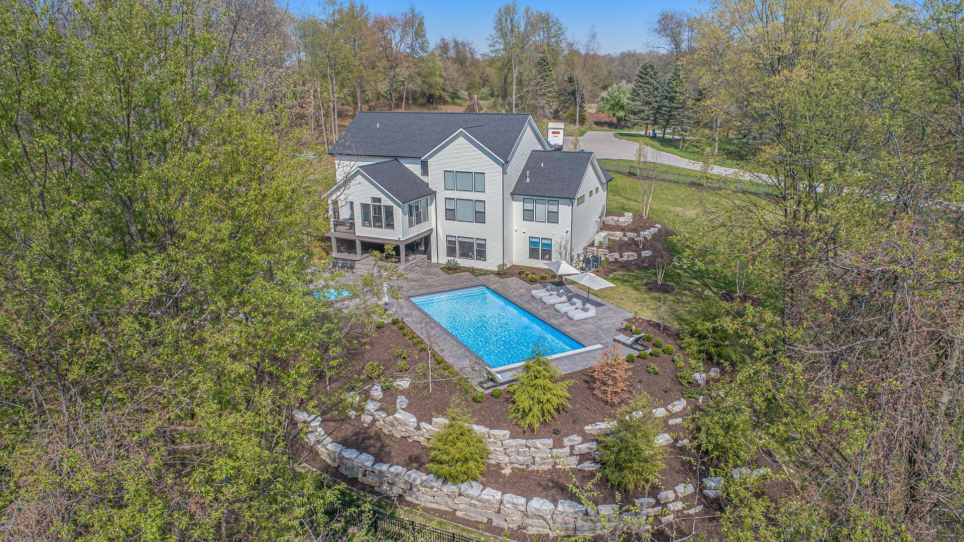 House with a pool surrounded by trees. Blue pool, gray house, stone retaining wall, and green foliage.