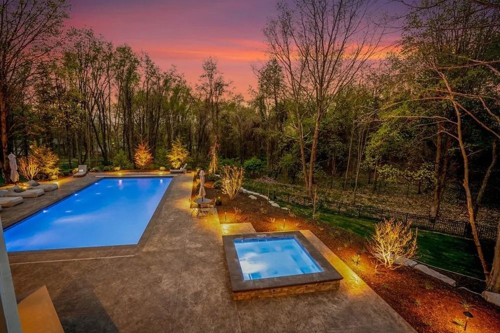 Pool and hot tub at dusk surrounded by trees, illuminated by ambient lighting.
