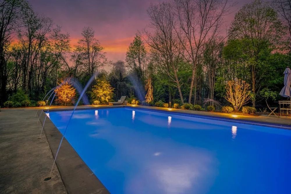 Swimming pool at dusk with water fountains, surrounded by trees and colorful sky.