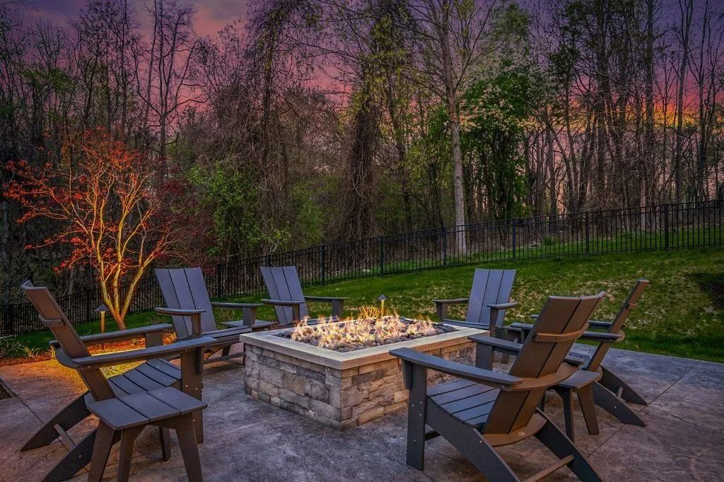 Fire pit with six gray Adirondack chairs in a backyard at dusk.