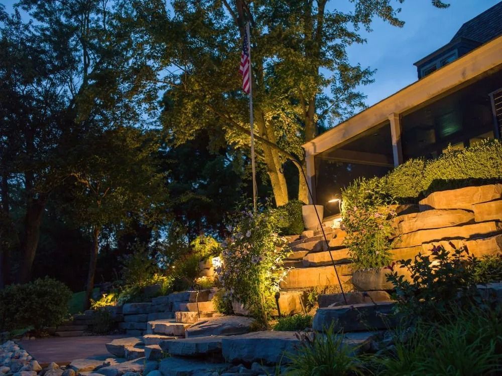 Outdoor scene with lit stone steps, a house, trees, and an American flag at dusk.