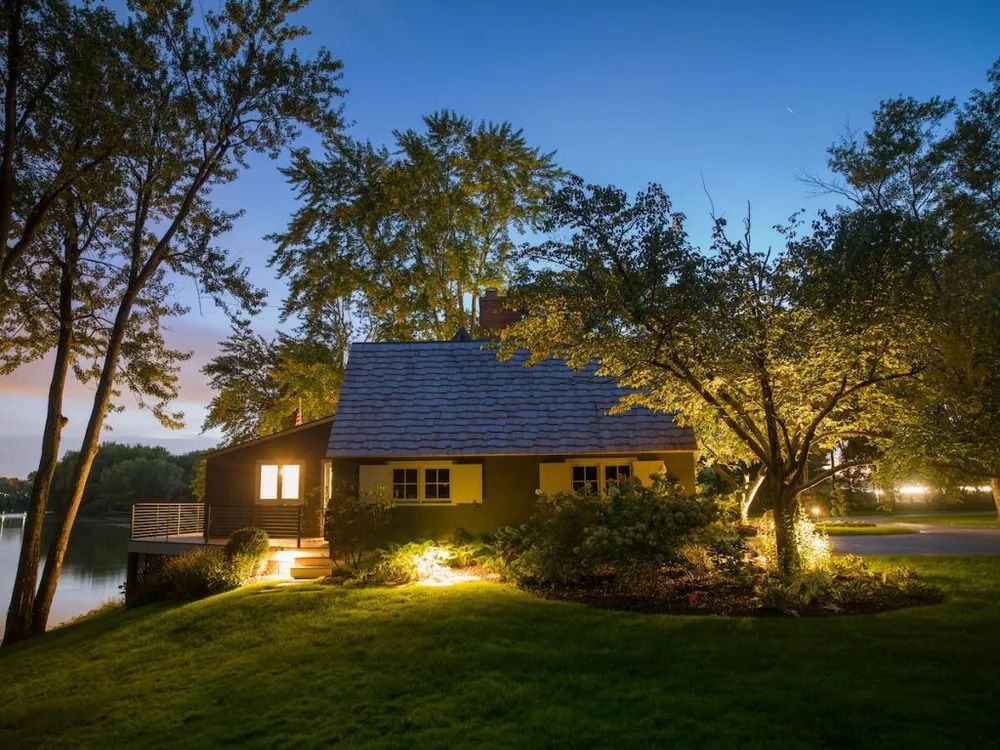Cottage illuminated at dusk near water, with trees and soft lighting on the lawn.