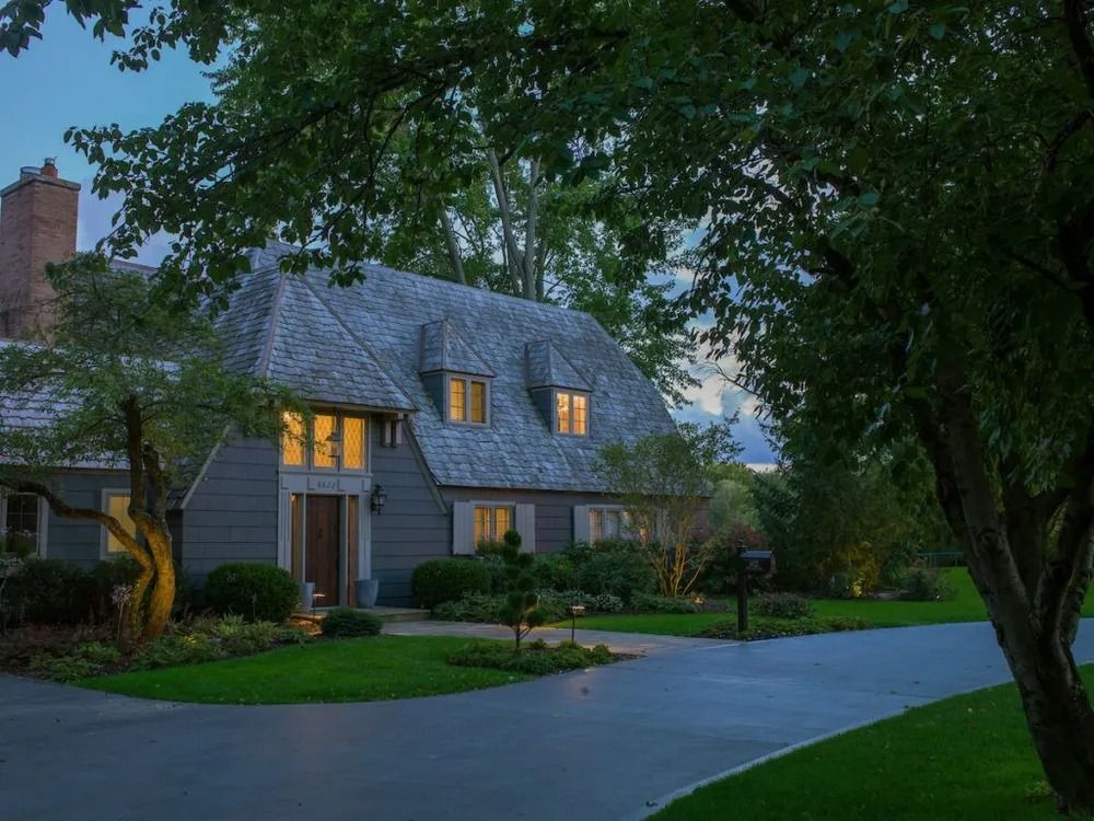 A gray house with a wood shingle roof illuminated by lights at dusk, with trees in the foreground.