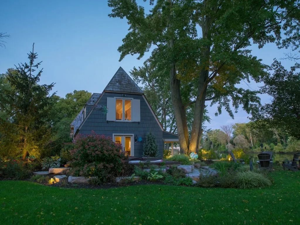 A-frame house with a gray exterior, set in a green yard with surrounding trees. Lit windows against the dusk sky.