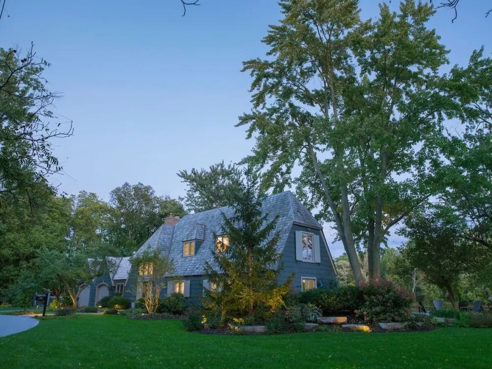 Blue house with a gray roof at dusk surrounded by trees and a green lawn.