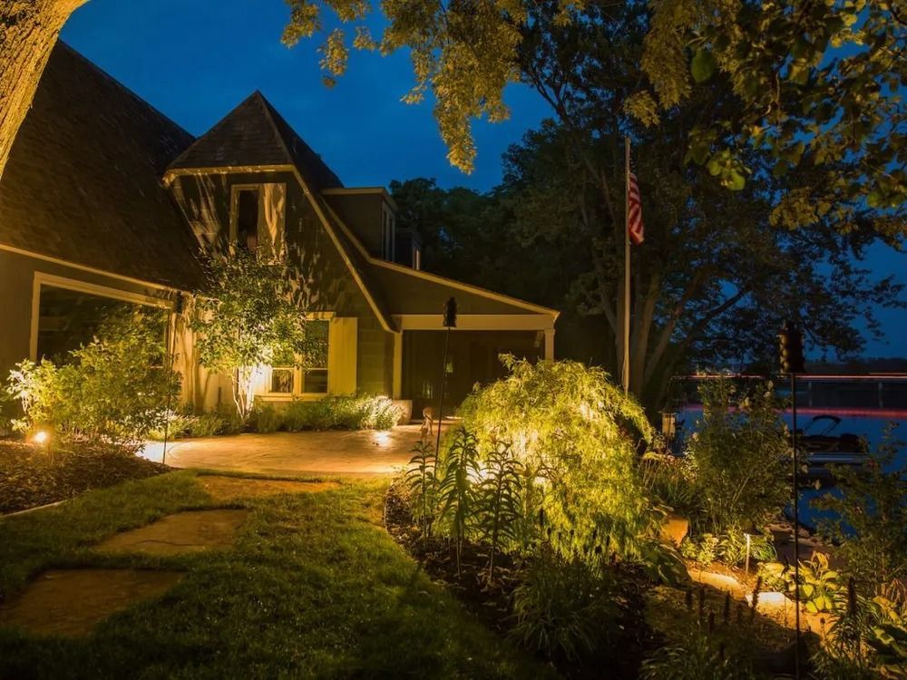 Nighttime view of a house with landscape lighting illuminating the front, walkway, and shrubs.