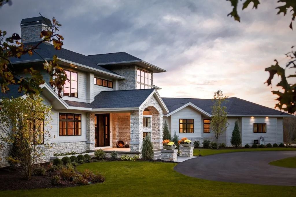 Large, two-story stone house with dark roof and circular driveway. Cloudy sky at dusk.