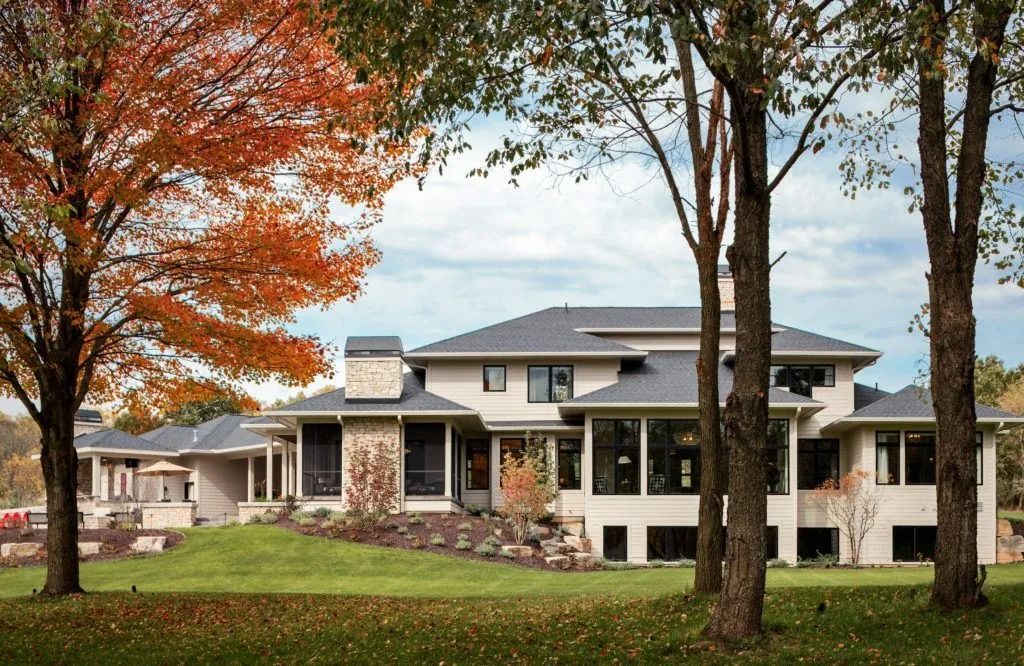 Modern two-story house with light-colored exterior and dark roof, surrounded by trees and a grassy lawn in autumn.