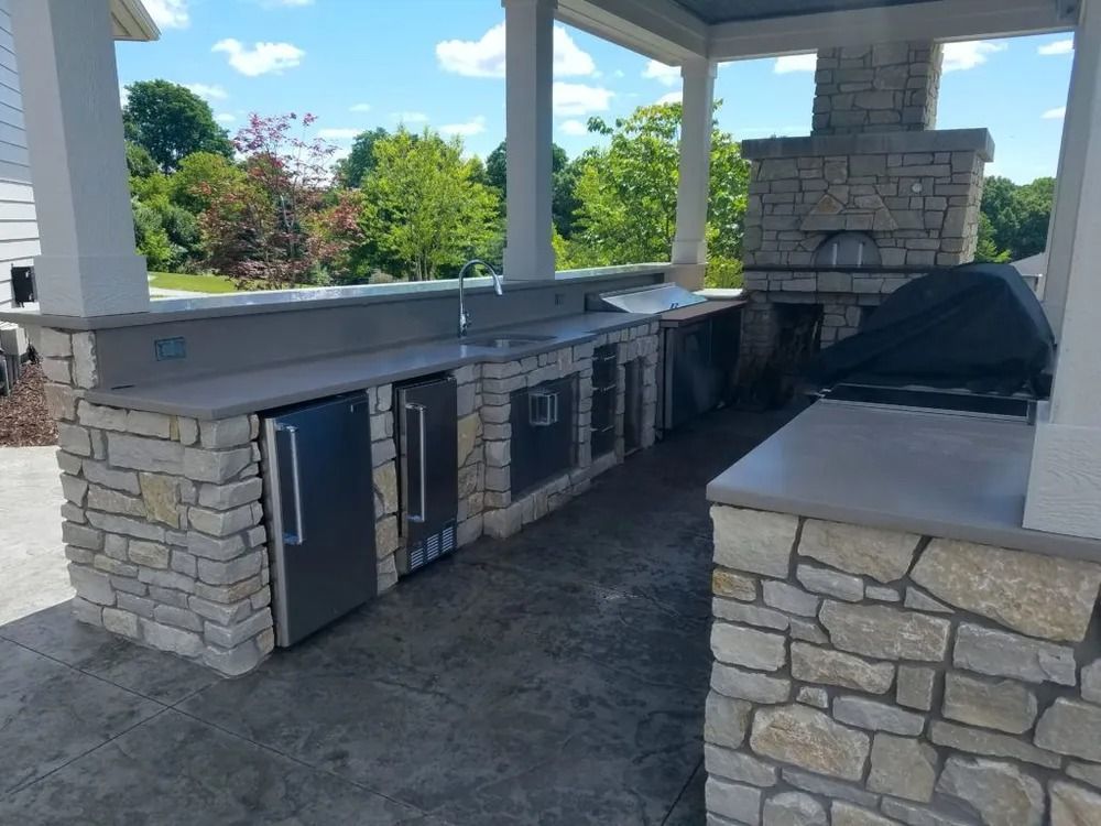 Outdoor kitchen with stone facade, stainless steel appliances, and pizza oven under a covered patio.