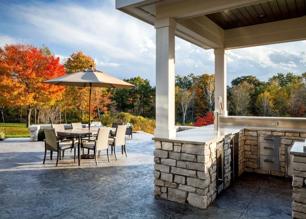 Outdoor patio with stone kitchen, dining set, and colorful autumn trees in the background.