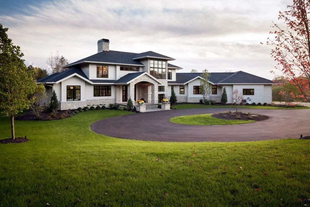 Luxurious white house with black roof and circular driveway. Lush green lawn under a cloudy sky.
