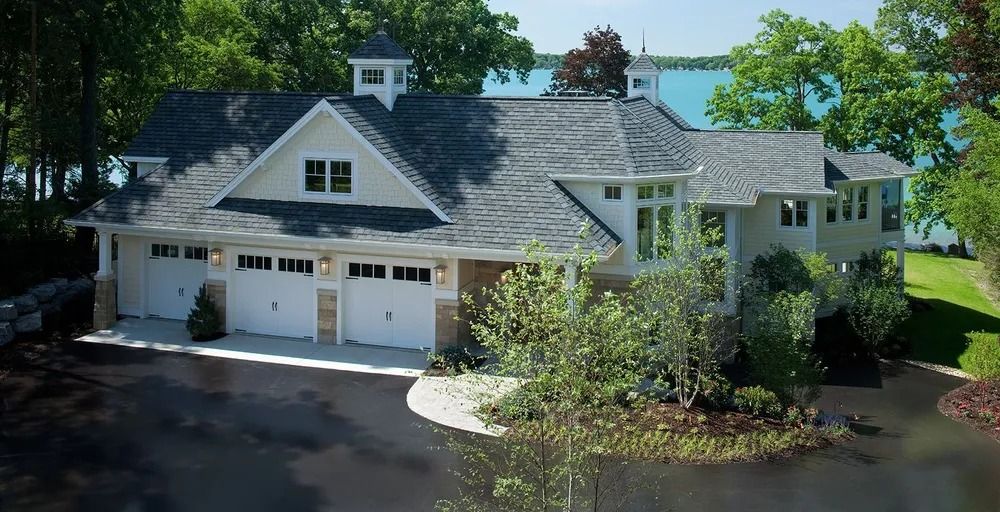 Beige and white lakeside house with three-car garage, dark gray roof, and lush green trees.