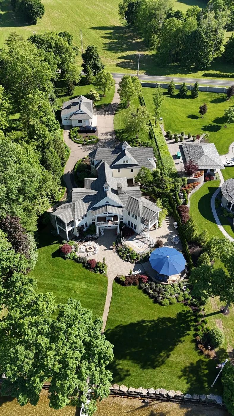 Aerial view of a white mansion with a blue umbrella on a large green lawn, surrounded by trees.