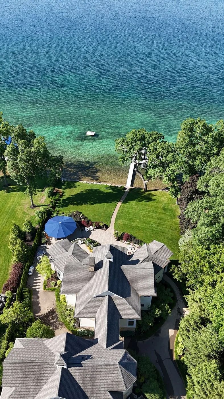 Overhead view of a house with a blue umbrella on a lawn leading to a dock on clear turquoise water.