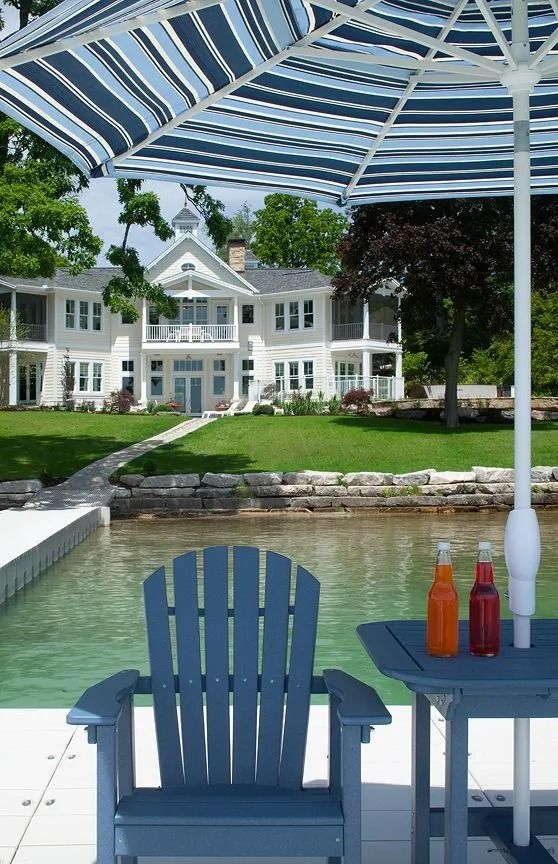 Blue Adirondack chair and table on a dock, overlooking a large white house and lake, under a striped umbrella.