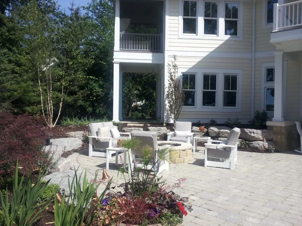 Patio with white chairs, fire pit, and stone retaining wall next to a light-colored house.