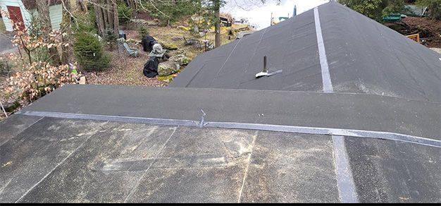 View of a dark, flat roof with a pointed section in the background. A black sealant tape runs along the edges.