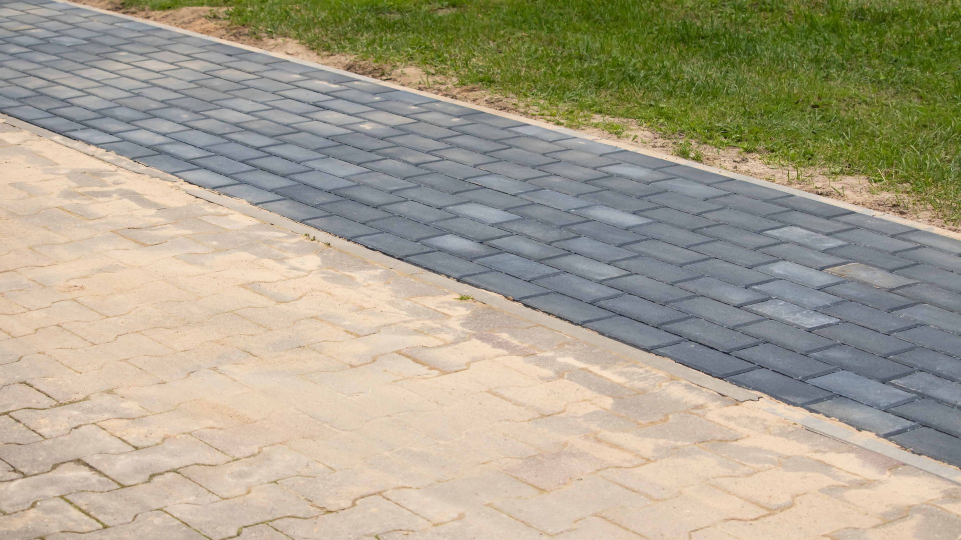 Paved walkway with gray brick pavers next to a sandy surface, bordered by grass.