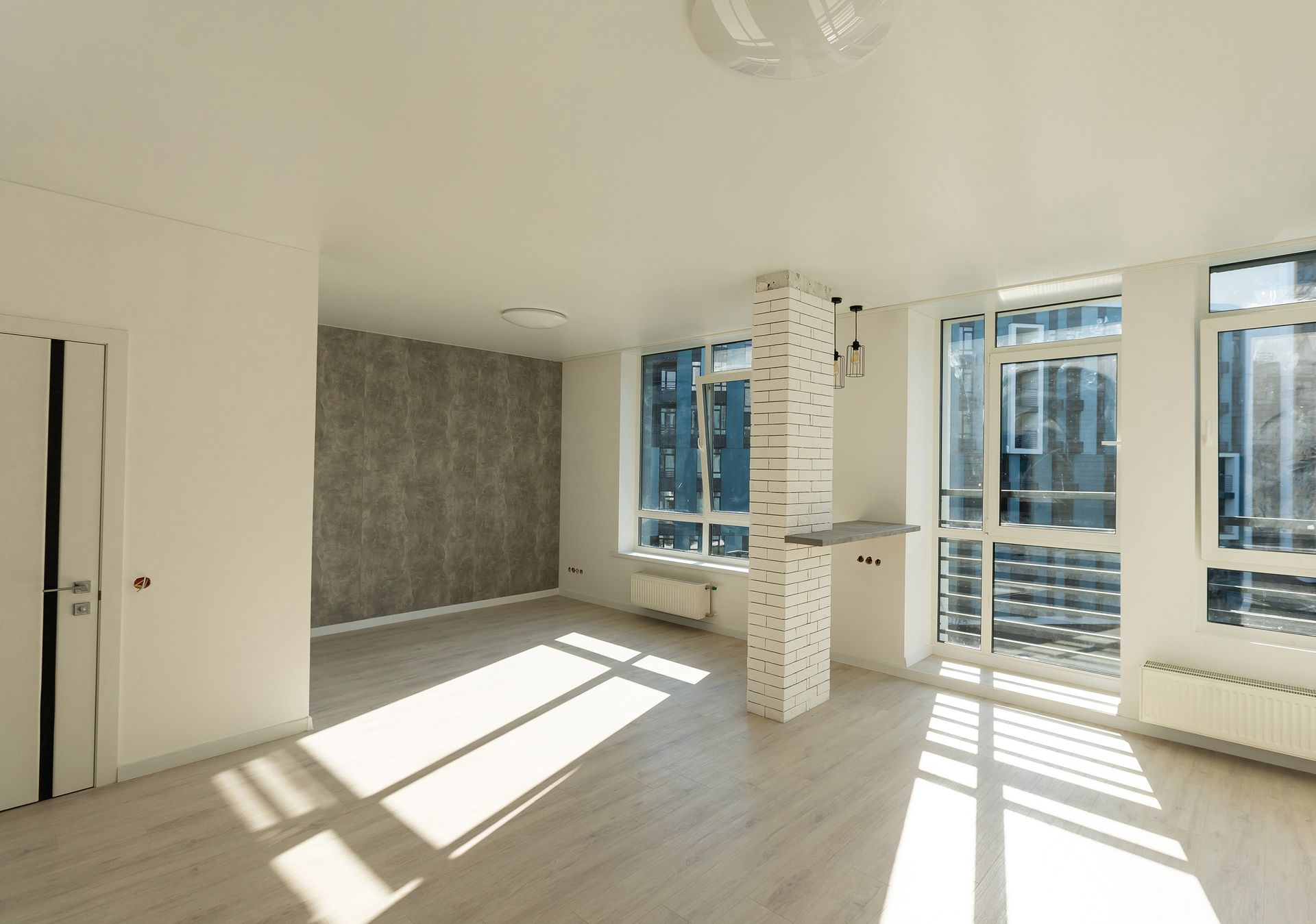 Empty, sunlit room with light wood floors, tall windows, and a partially textured accent wall.