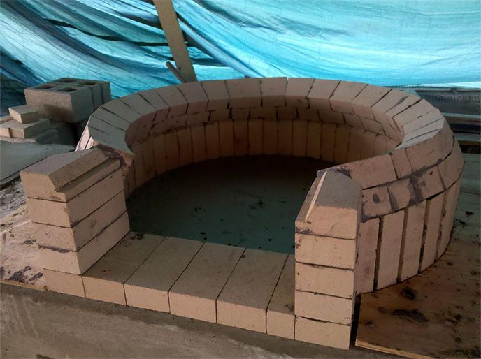 Brick oven under construction, circular design with front opening, light brown bricks, blue tarp background.