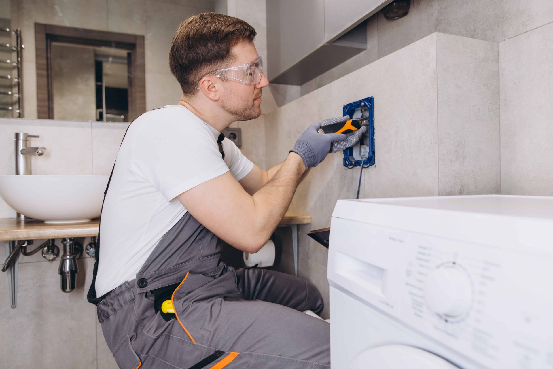 Man in gray coveralls and safety glasses installing electrical outlet near a washing machine.