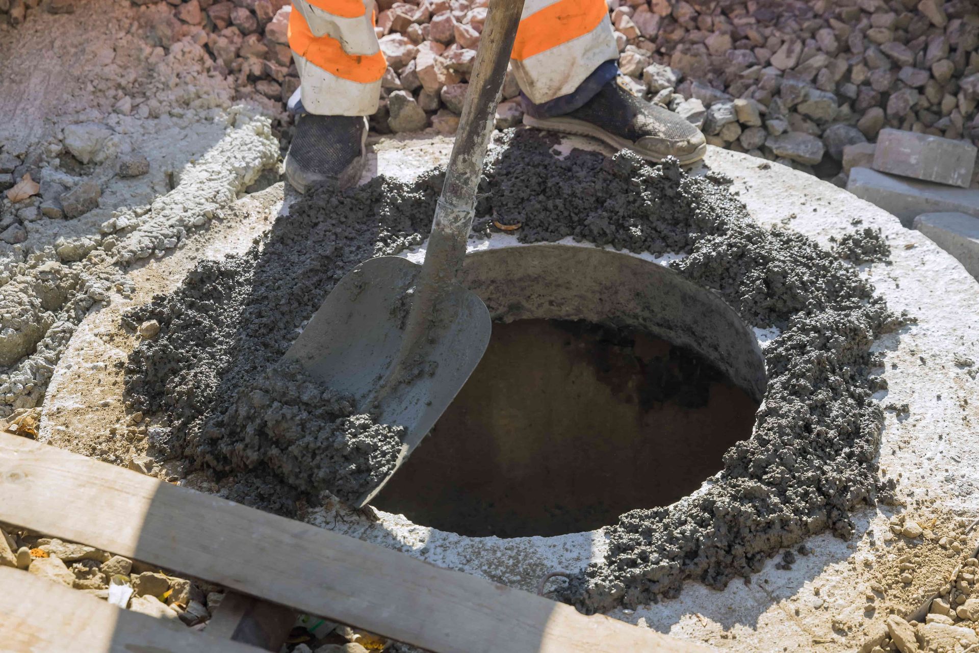 Person in orange work pants mixing wet cement with a shovel on a construction site.
