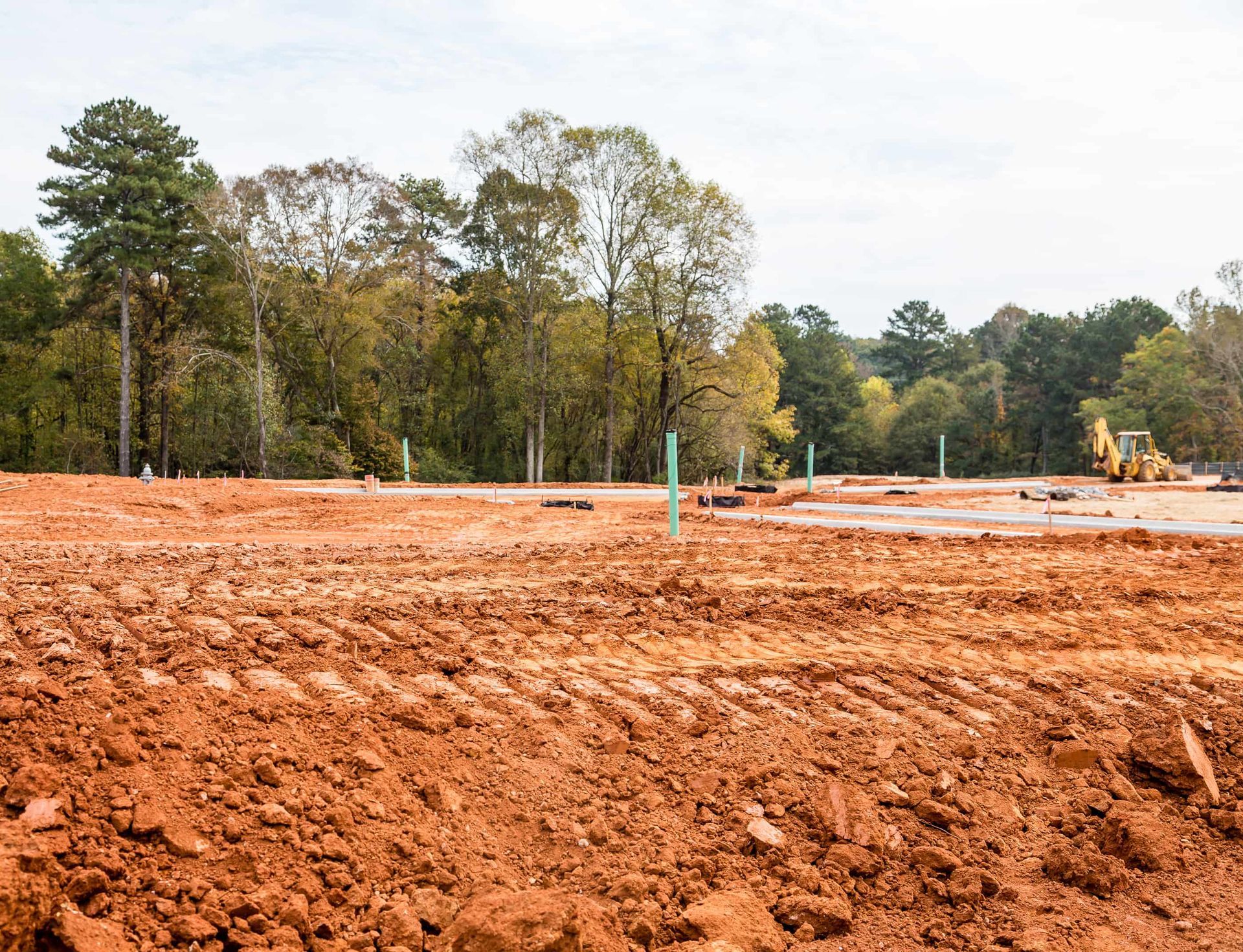 Construction site with red soil, tire tracks, and a yellow excavator. Trees in the background.