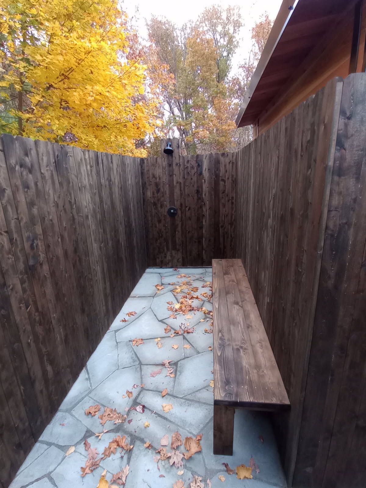 Outdoor shower with wooden walls, stone floor, and a bench; autumn leaves and trees visible.