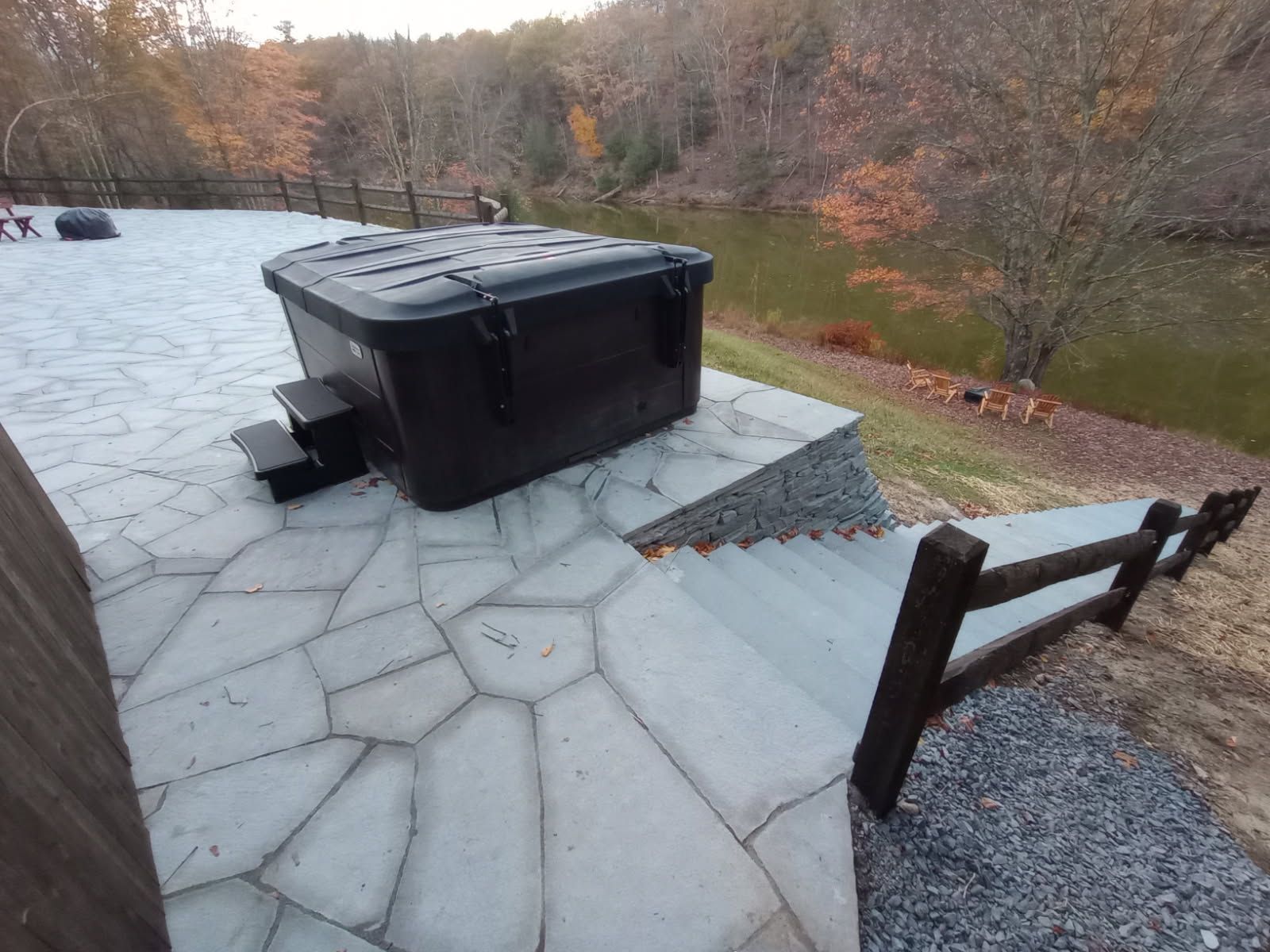 Black hot tub with steps on a stone patio overlooking a lake, bordered by a wooden fence.