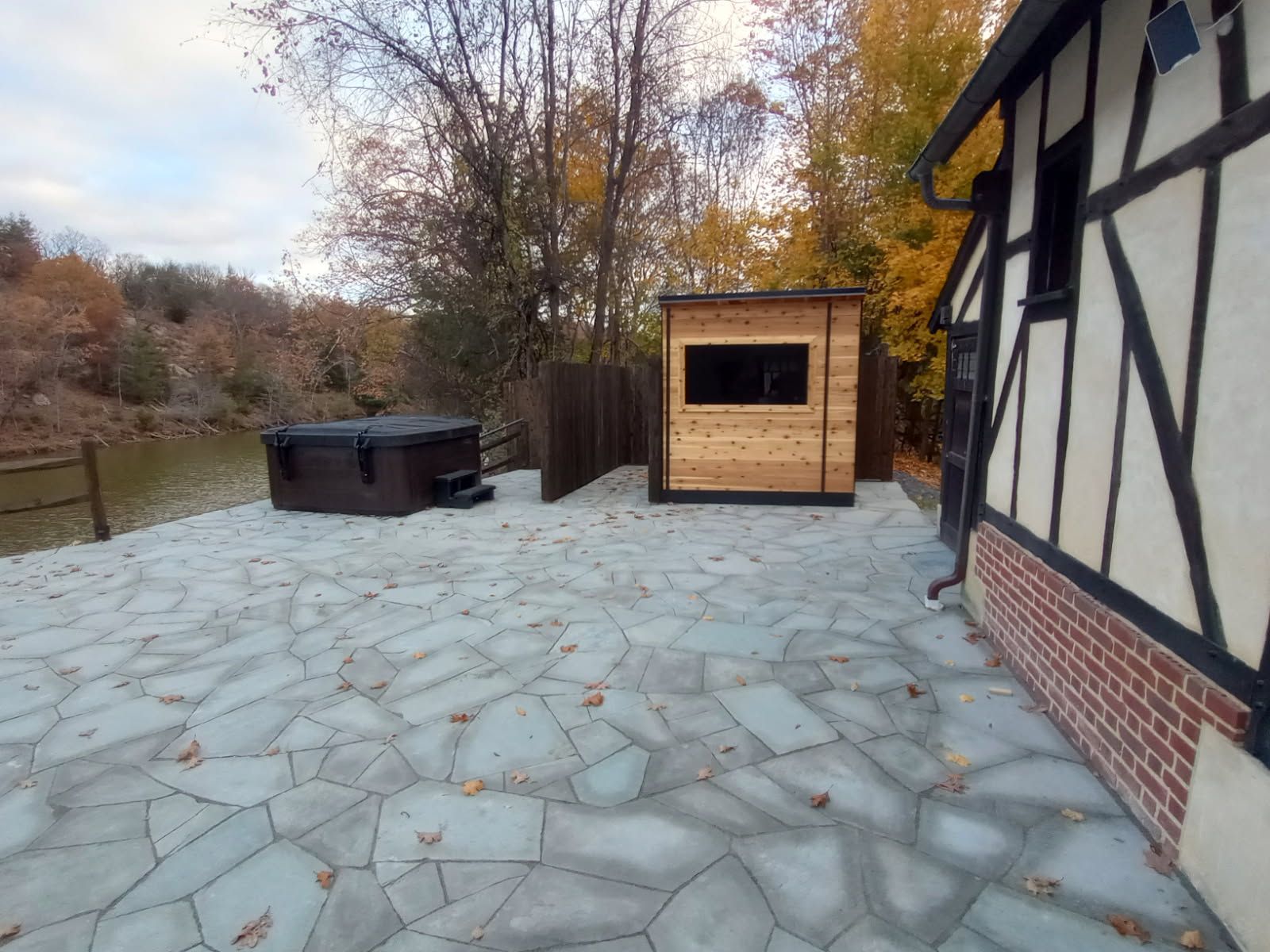 Hot tub, wooden shed, and house with brickwork on a stone patio by a lake.
