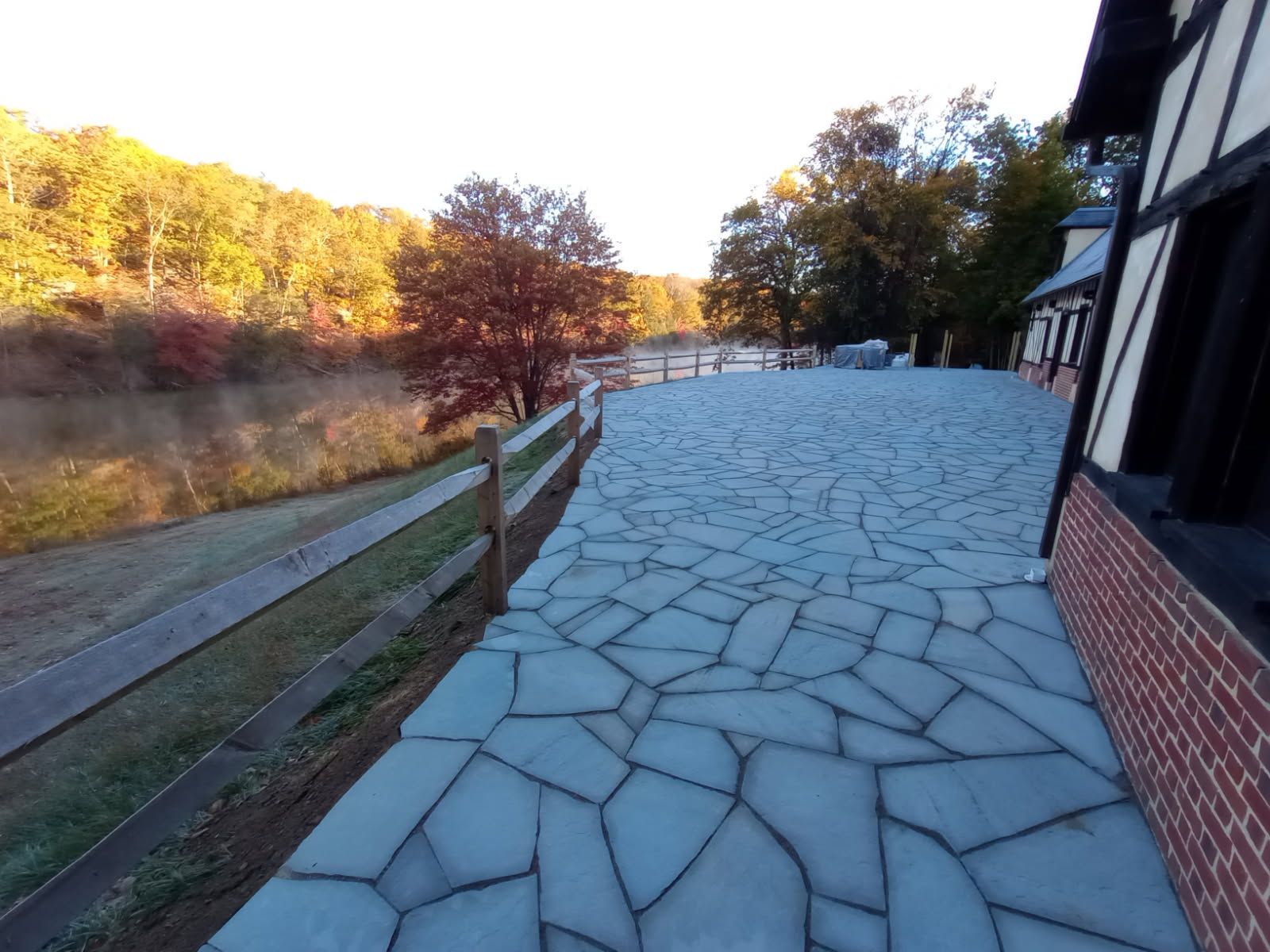 Stone patio next to a wooden fence and river, Tudor-style building on the right, autumn foliage in the background.