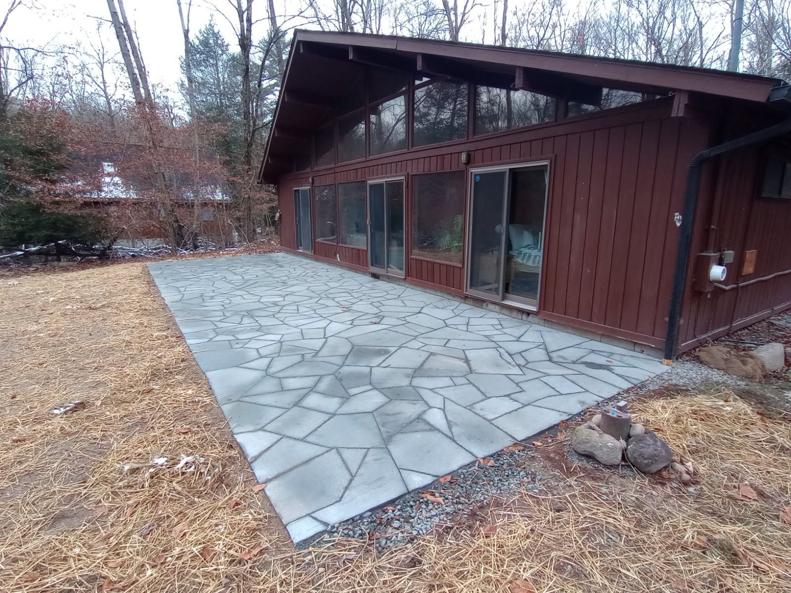 A brown cabin with a stone patio. The patio is surrounded by dry grass and trees.
