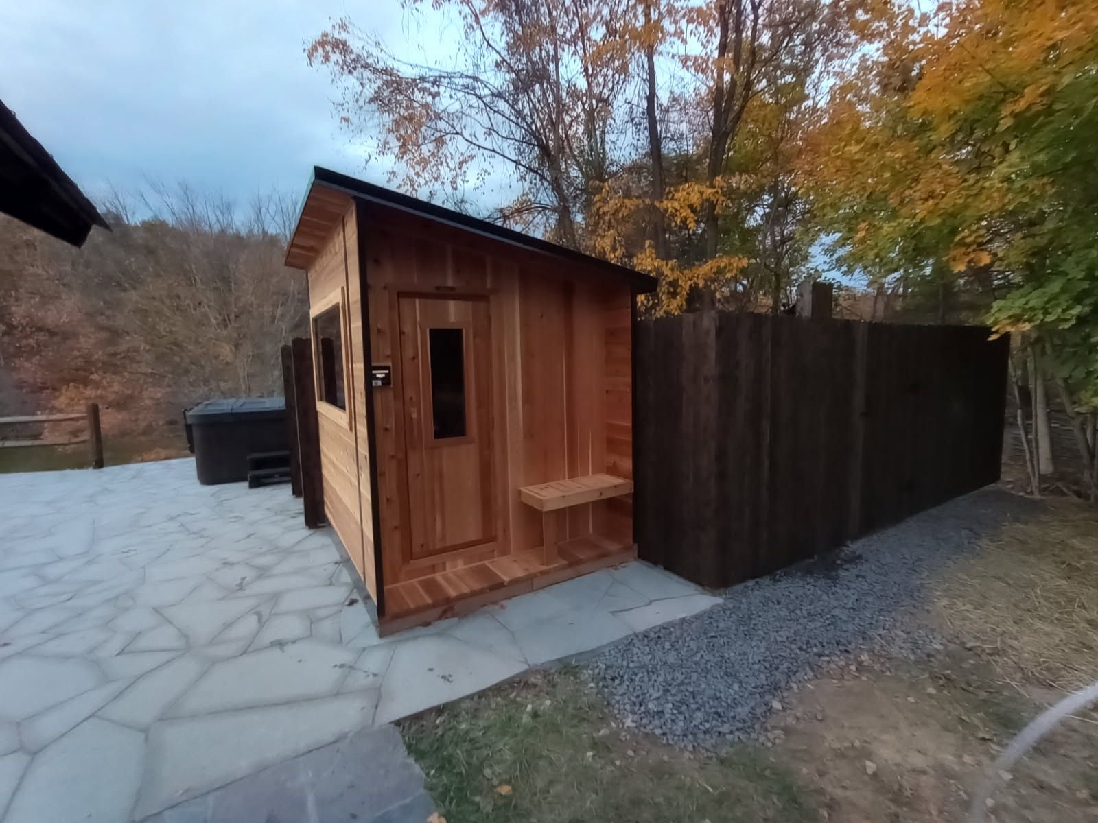 Wooden sauna building with bench and small window next to a dark wooden fence and stone patio.