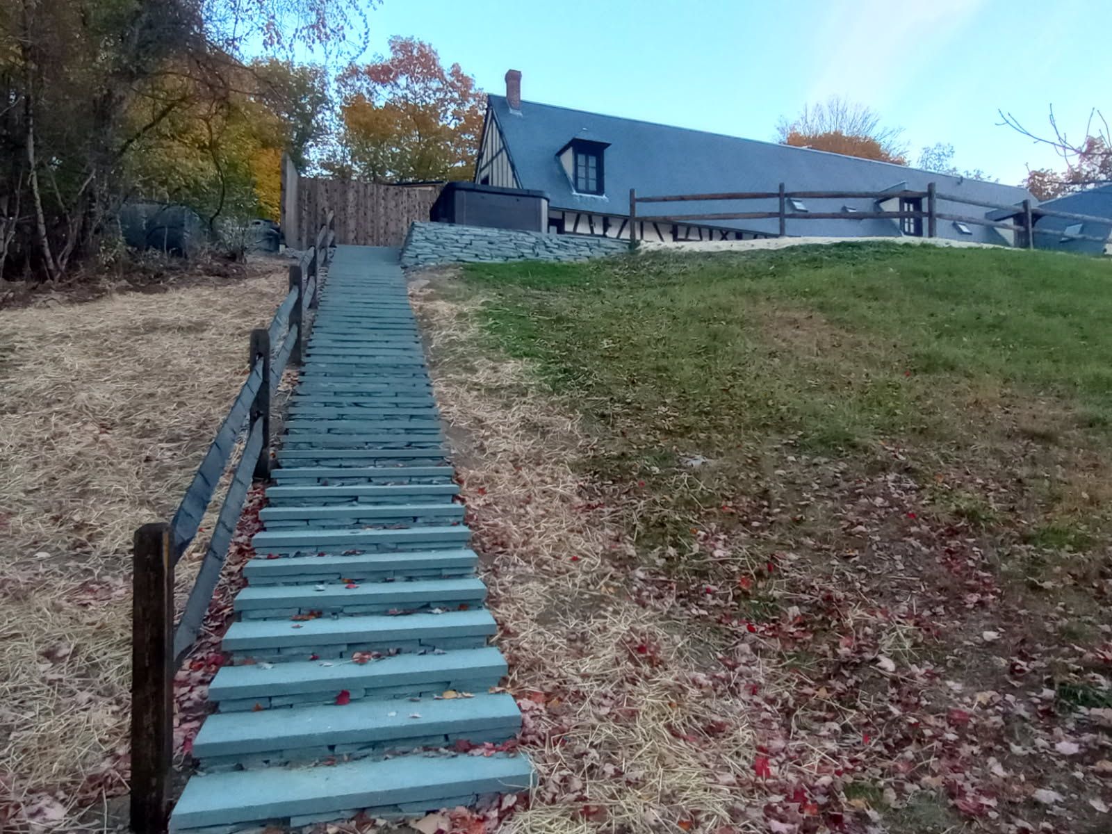 Outdoor staircase leading uphill towards a house with a sloped roof. Autumn leaves and grass visible.