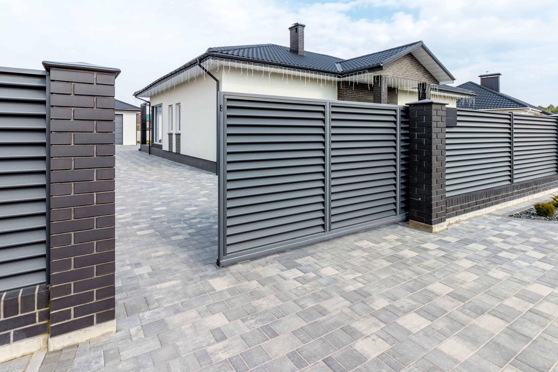 Gray louvered gate and fence with brick posts in front of a modern white house and paved driveway.