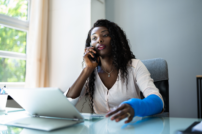 A man with a broken arm is sitting at a desk with a laptop and talking on a phone.