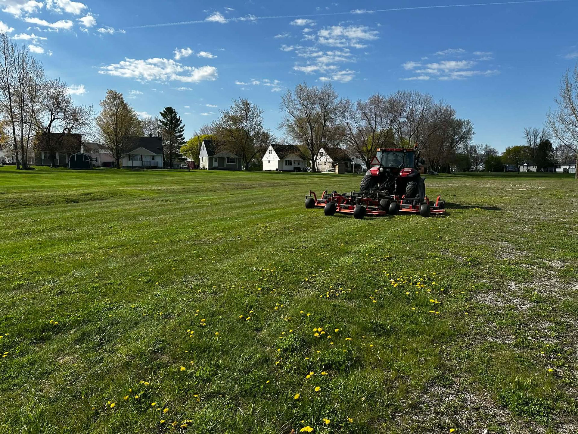 Red mower mowing a grassy field under a blue sky with houses and trees in the background