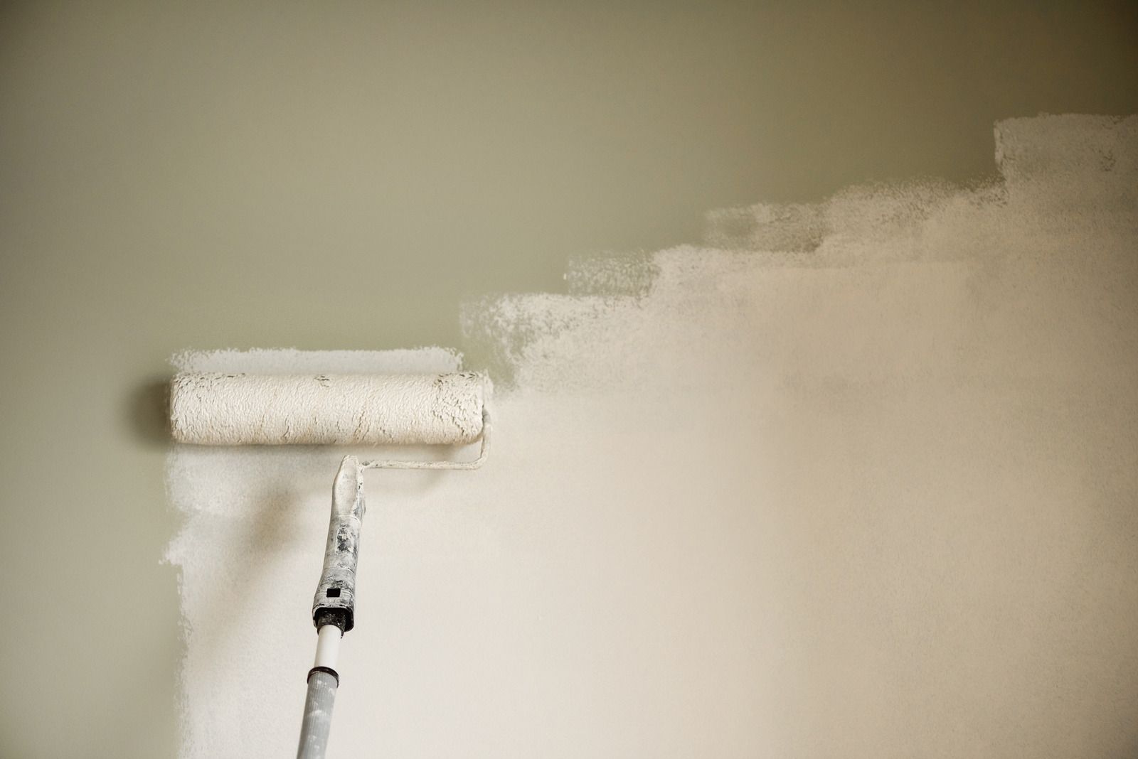 A paint roller applying white paint to a wall, covering a lighter green color.