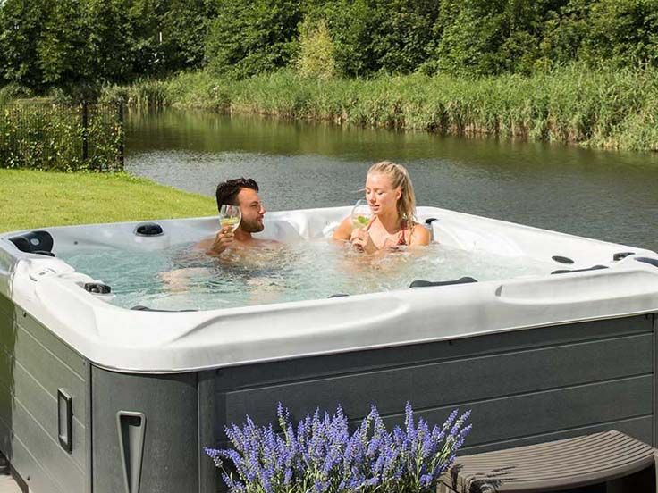 Couple in hot tub, drinking wine, overlooking a calm lake with green vegetation.