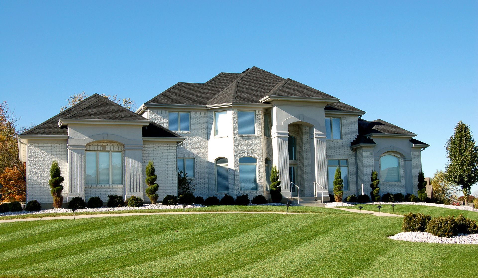 Large white luxury house with a dark roof and manicured green lawn under a clear blue sky