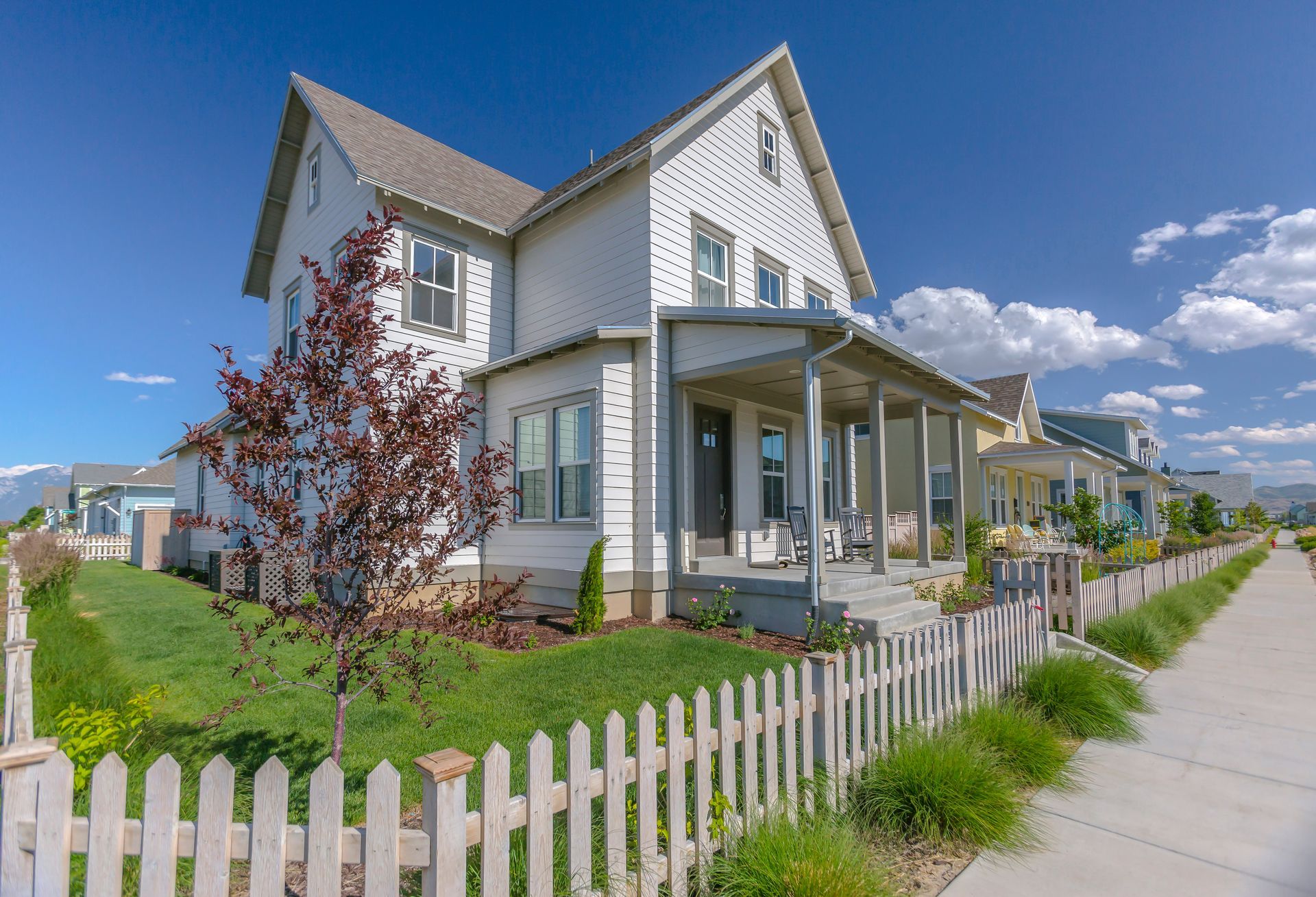 White two-story house with porch and picket fence on a sunny suburban street