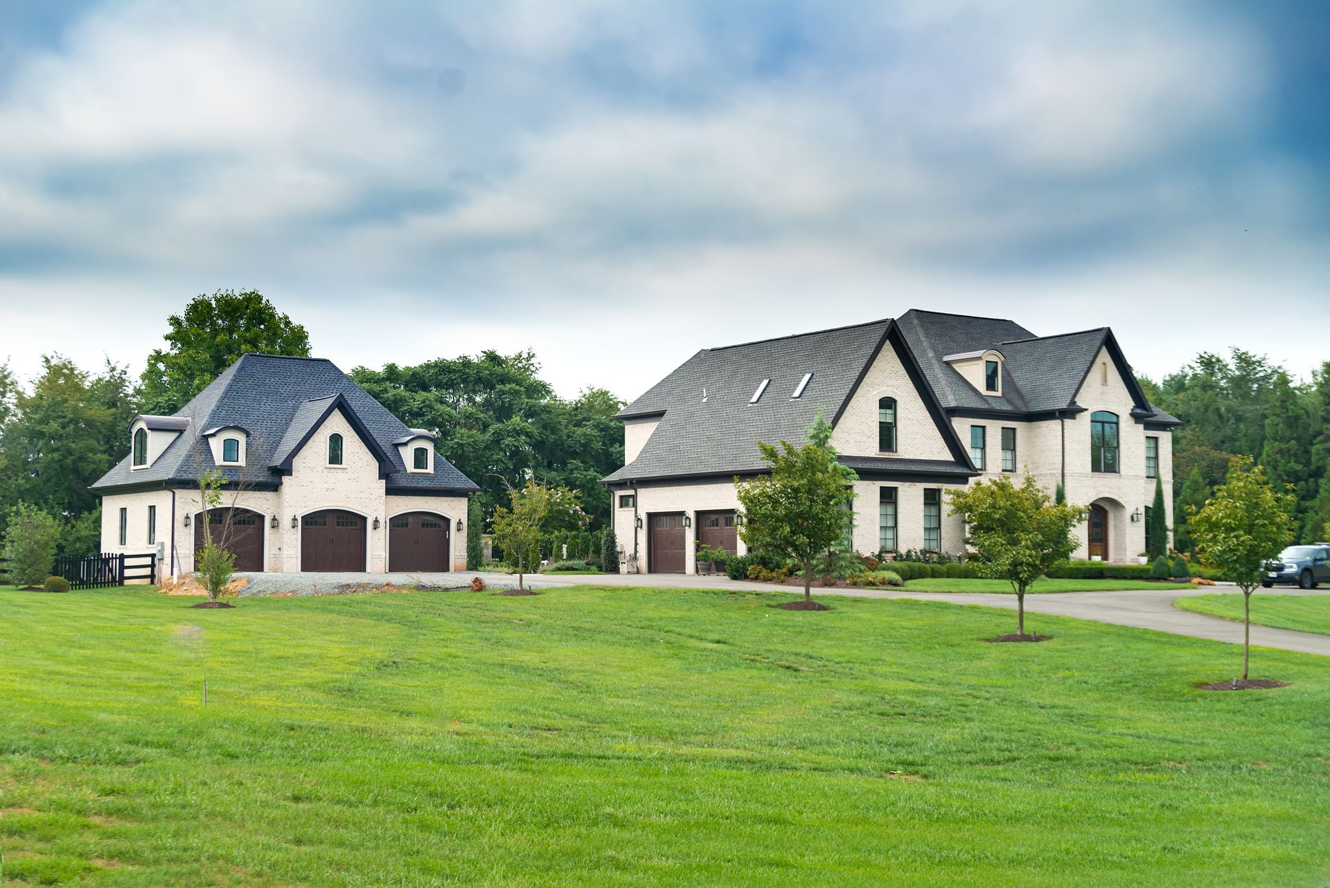Two white suburban houses with garages beside a green lawn under a cloudy sky