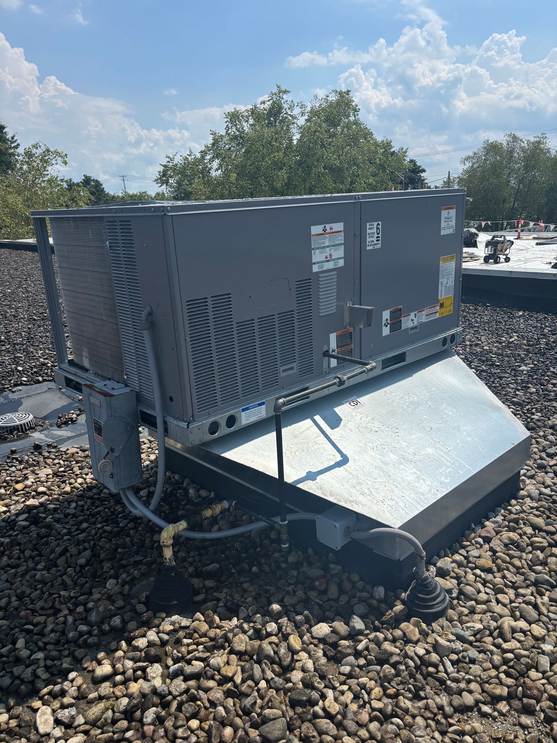 Rooftop air conditioning unit on a gravel-covered roof with blue sky and trees in the background.